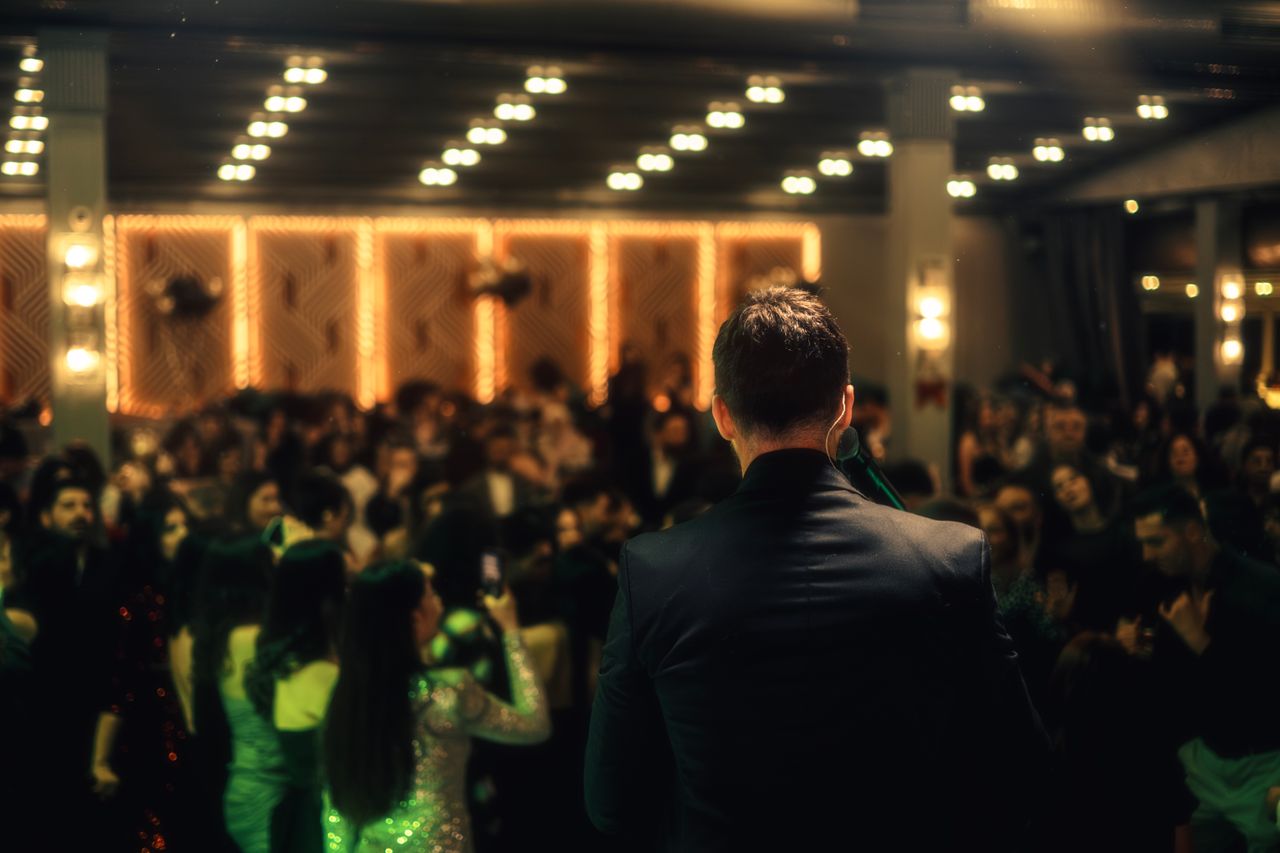 The Art of Drawing Readers In: Your attractive post title goes here A crowded indoor night event with a man addressing the audience, vibrant lighting enhances the scene.